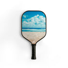 A pickleball paddle with a black handle and a printed beach scene on the face, featuring a blue sky with white clouds, ocean waves, and pale sand. The brand name 'Ehakai' is visible near the throat of the paddle.