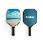 Two Elakai pickleball paddles shown side by side on a white background. The paddle on the left features a beach and ocean print, while the paddle on the right is solid blue with the white 'Elakai' brand logo.