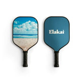 Two Elakai pickleball paddles shown side by side on a white background. The paddle on the left features a beach and ocean print, while the paddle on the right is solid blue with the white 'Elakai' brand logo.