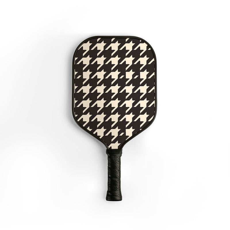 A pickleball paddle featuring a classic black and cream houndstooth pattern on the paddle face, with a textured black grip handle, shown against a plain white background.