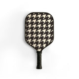 A pickleball paddle featuring a classic black and cream houndstooth pattern on the paddle face, with a textured black grip handle, shown against a plain white background.