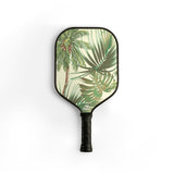 A pickleball paddle with a tropical palm tree print on a cream background, featuring a black frame and a black textured grip handle, shown against a plain white background.