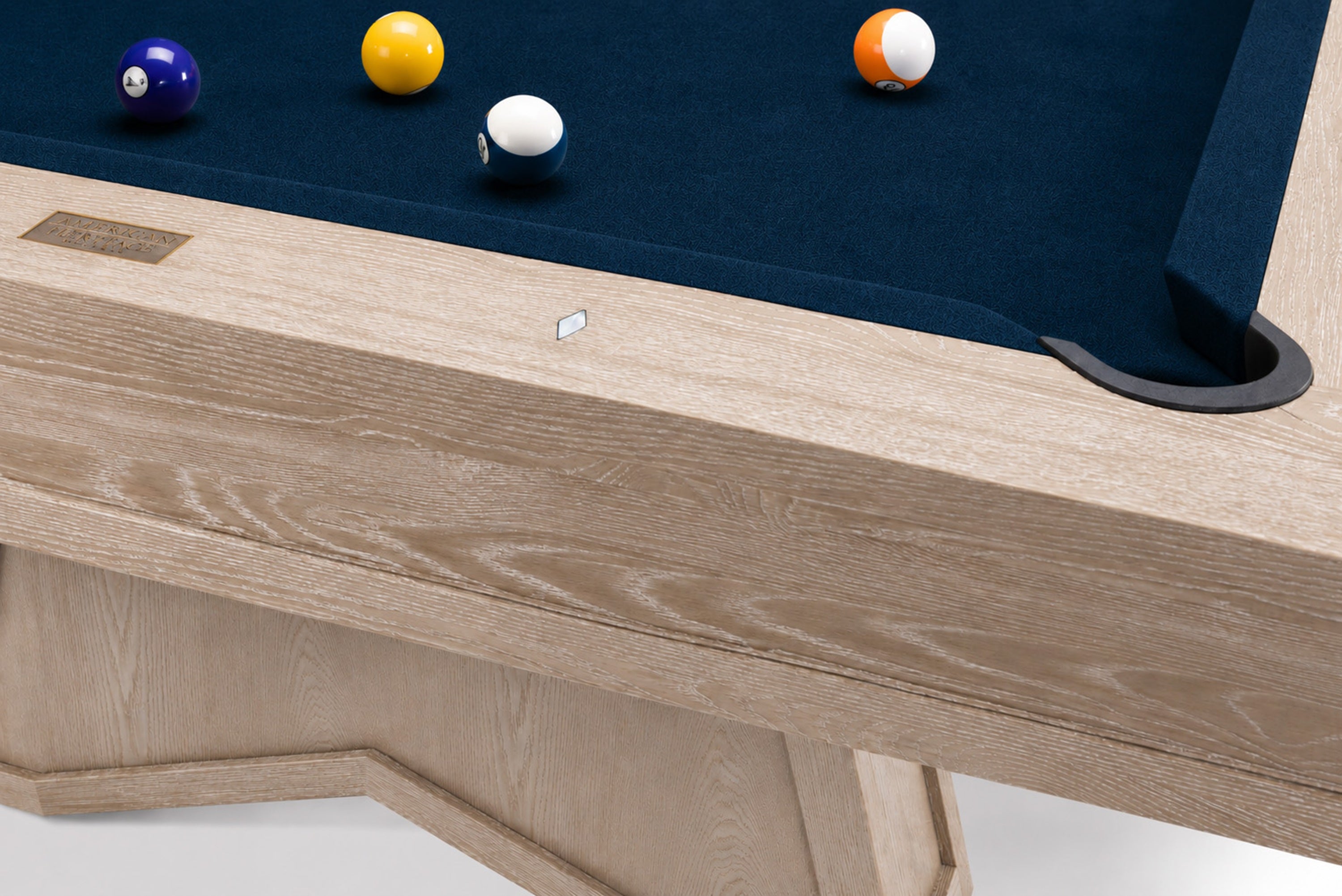 Close-up of a pool table corner showing a light natural wood frame, dark blue felt cloth, and several billiard balls including a yellow 8-ball, white cue ball, and orange ball.