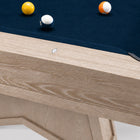 Close-up of a pool table corner showing a light natural wood frame, dark blue felt cloth, and several billiard balls including a yellow 8-ball, white cue ball, and orange ball.