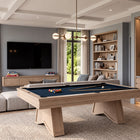 A modern living room featuring a light wood pool table with a dark blue felt surface, styled in a high-end contemporary space with a sectional sofa, built-in shelving, and a bar area.
