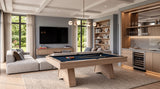 A modern living room featuring a light wood pool table with a dark blue felt surface, styled in a high-end contemporary space with a sectional sofa, built-in shelving, and a bar area.