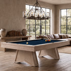 A light wood pool table with a deep blue felt surface is shown in a modern, sunlit living room with large windows, a sectional sofa, and a chandelier.