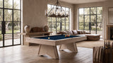 A light wood pool table with a deep blue felt surface is shown in a modern, sunlit living room with large windows, a sectional sofa, and a chandelier.