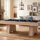 A light natural wood pool table with a blue felt playing surface and colorful billiard balls, set in a beautifully styled living room with a fireplace, sofa, and framed wall art.