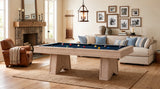 A light natural wood pool table with a blue felt playing surface and colorful billiard balls, set in a beautifully styled living room with a fireplace, sofa, and framed wall art.