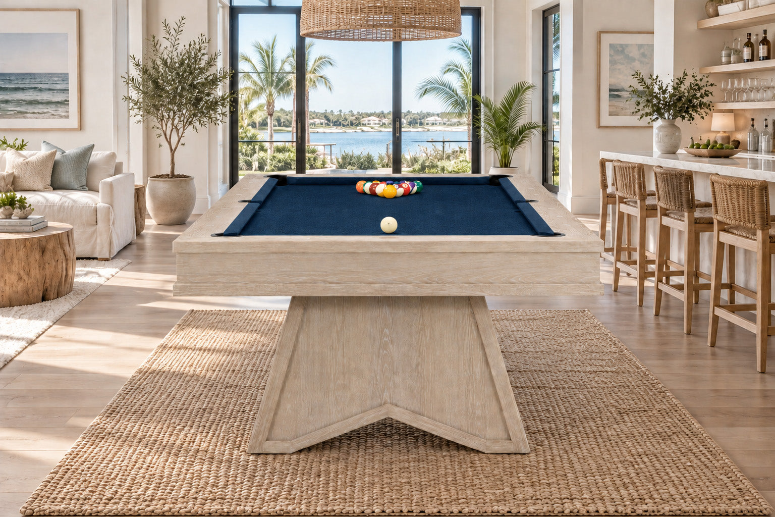A light natural wood pool table with a dark blue felt surface, set in a bright, modern beach-themed living room with a large woven jute rug.