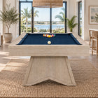 A light natural wood pool table with a dark blue felt surface, set in a bright, modern beach-themed living room with a large woven jute rug.
