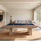 A modern wooden pool table with a blue felt surface is centered in a bright, contemporary room with large glass doors, a woven rug, and a light gray sofa in the background.