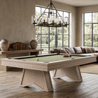 A light natural wood pool table with a green felt surface is shown in a modern, sunlit living room with large windows, a sectional sofa, and a chandelier.