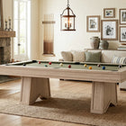 A light natural wood pool table with a green felt playing surface and scattered billiard balls, set in a styled living room with a fireplace, sofa, and framed wall art.