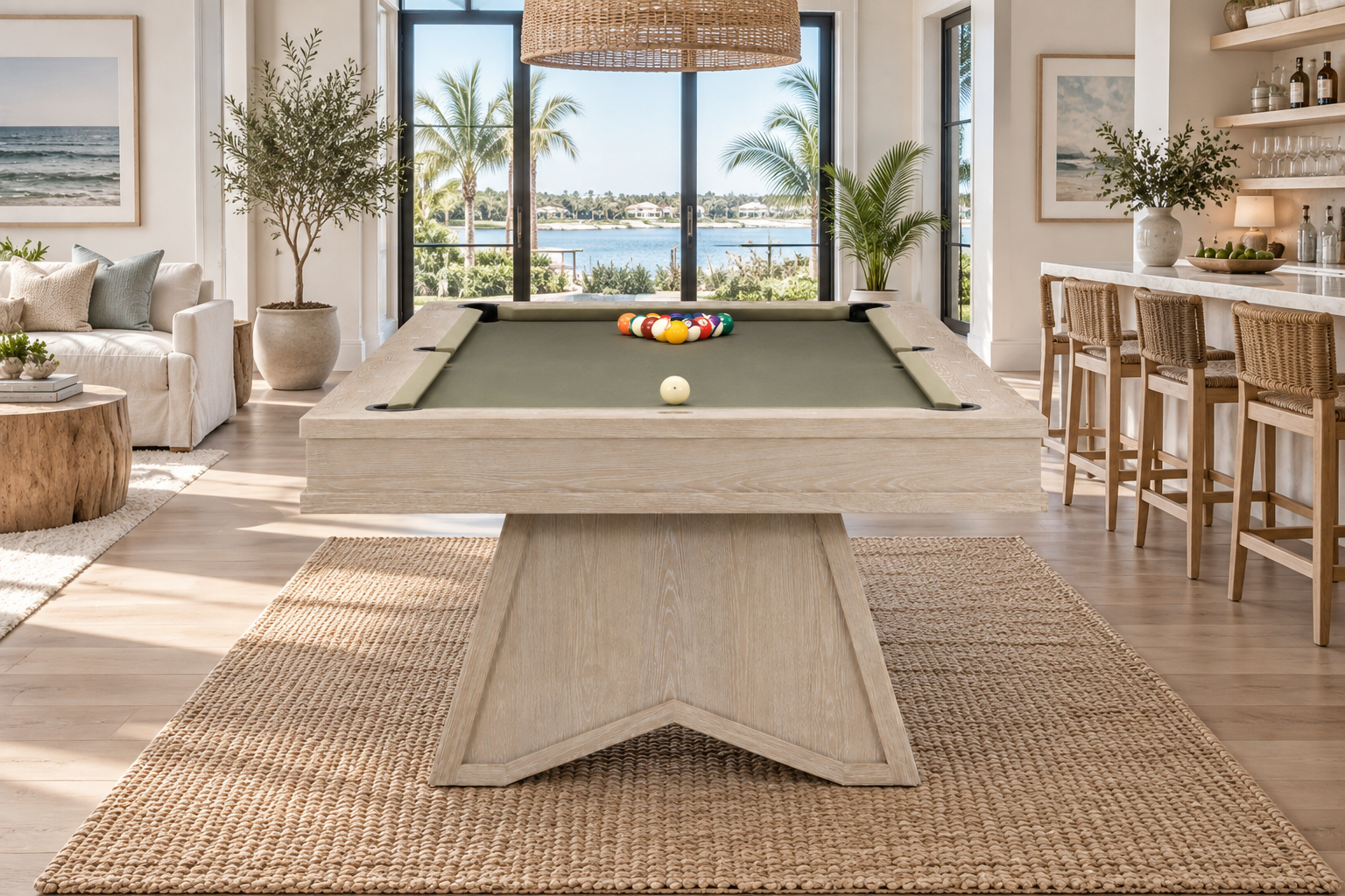 A light natural wood pool table with green felt sits in a bright, modern beach-view living room. The table is set on a large woven jute rug, with a rack of colorful billiard balls and a white cue ball on the green felt.