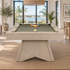 A light natural wood pool table with green felt sits in a bright, modern beach-view living room. The table is set on a large woven jute rug, with a rack of colorful billiard balls and a white cue ball on the green felt.