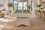 A light natural wood pool table with green felt sits in a bright, modern beach-view living room. The table is set on a large woven jute rug, with a rack of colorful billiard balls and a white cue ball on the green felt.