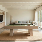A modern wooden pool table with a green felt surface is centered in a bright, contemporary room with large glass doors, a light wood TV wall, and a woven rug underneath.
