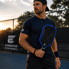 A man on a pickleball court at sunset wearing a navy Engage Pickleball shirt, black shorts, and a black cap, holding a black and blue Engage pickleball paddle.
