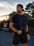A man on a pickleball court at sunset wearing a navy Engage Pickleball shirt, black shorts, and a black cap, holding a black and blue Engage pickleball paddle.