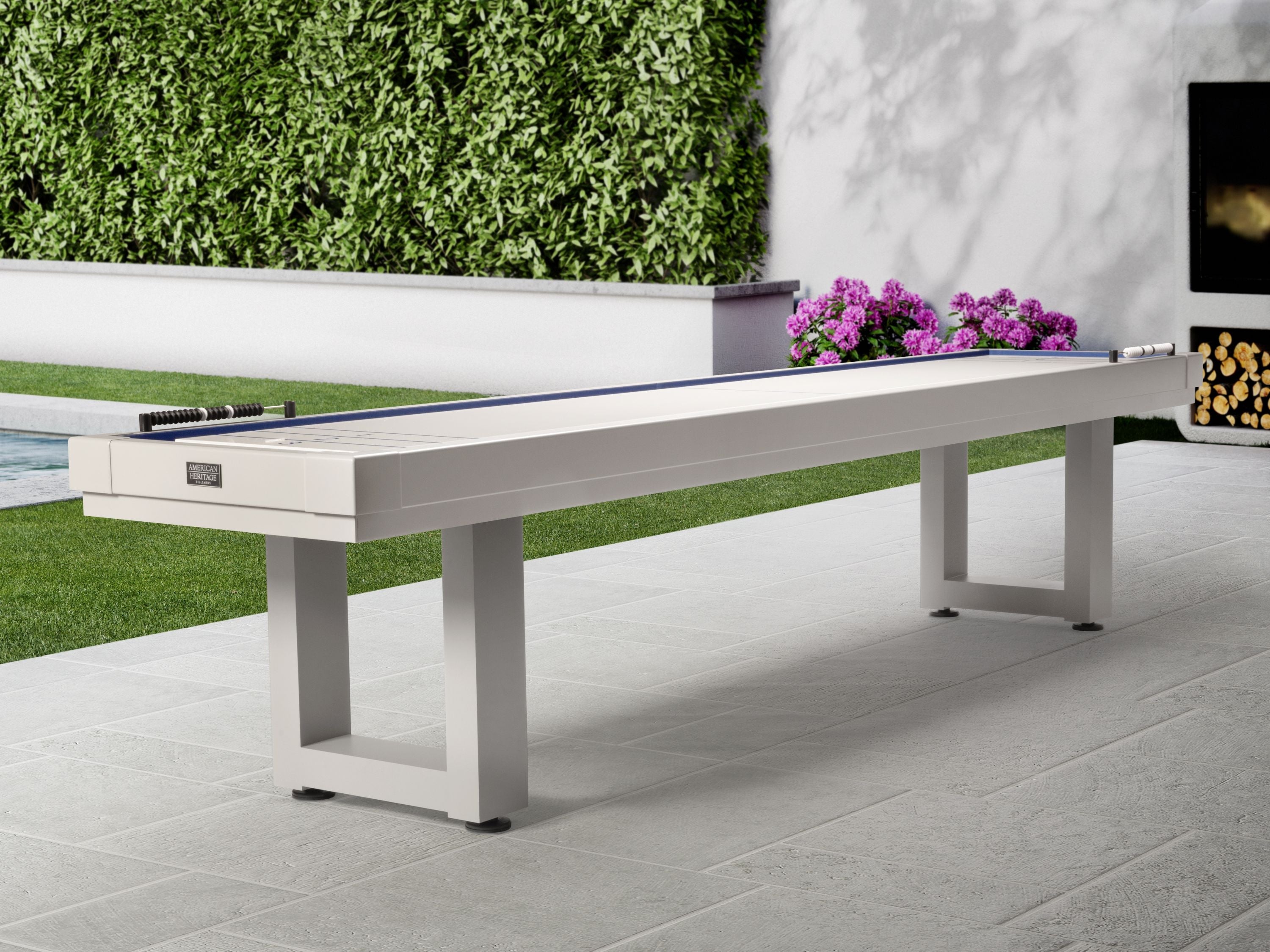 An American Heritage Lanai 12' Outdoor Shuffleboard Table in Pearl White sits near flowers.