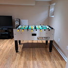 A Tornado Tournament Competition T-3000 Foosball Table in a basement with wood floors and a TV.