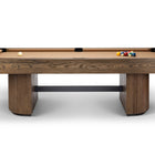 A modern pool table with a medium brown wood grain frame and legs, featuring a tan felt playing surface, a white cue ball, and a rack of colorful billiard balls, shown against a plain white background.