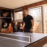 Family playing ping pong in a home setting with large windows.