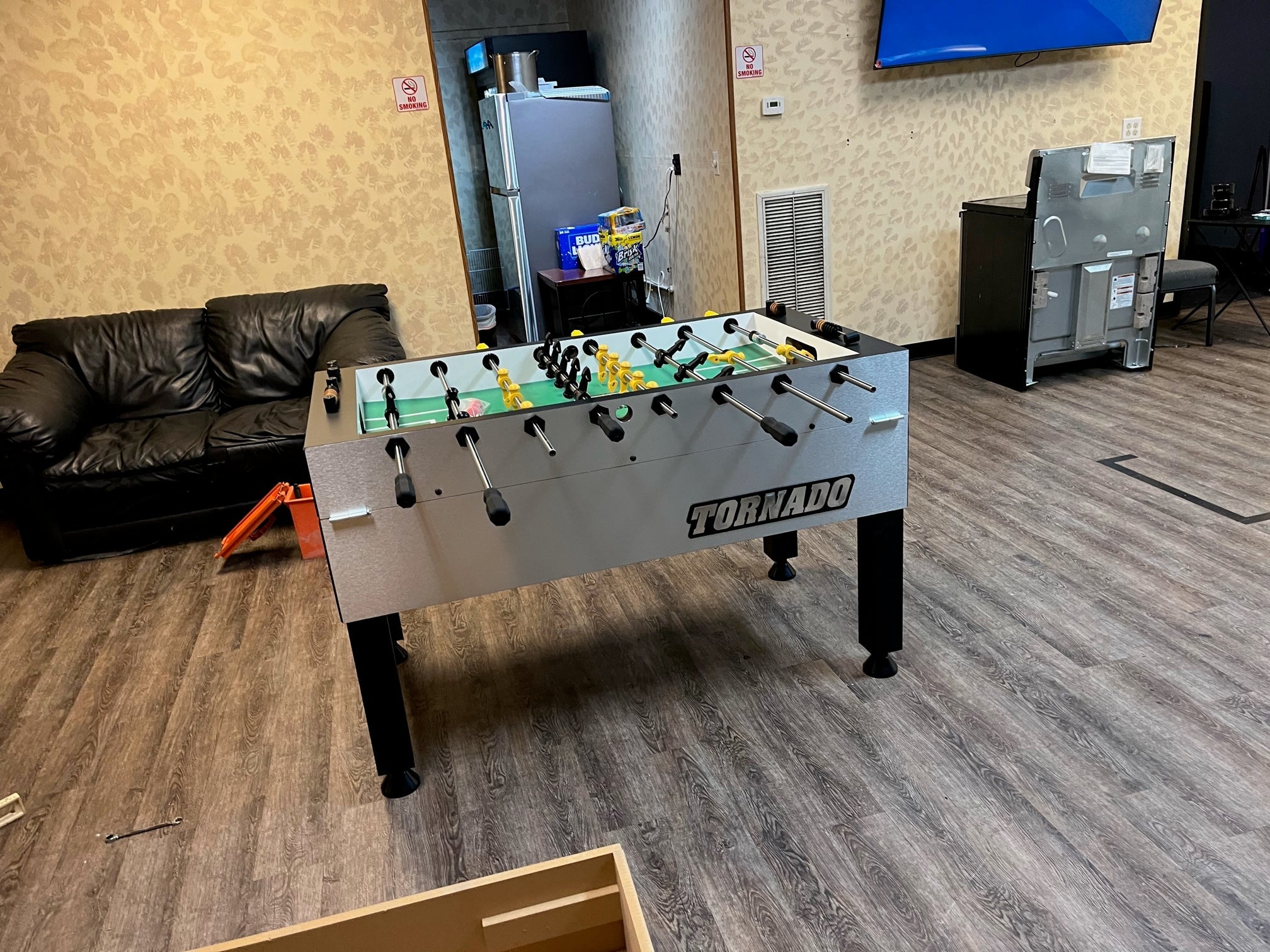 A Tornado T-3000 Professional Foosball Table in silver sits near a black couch and vending machines.