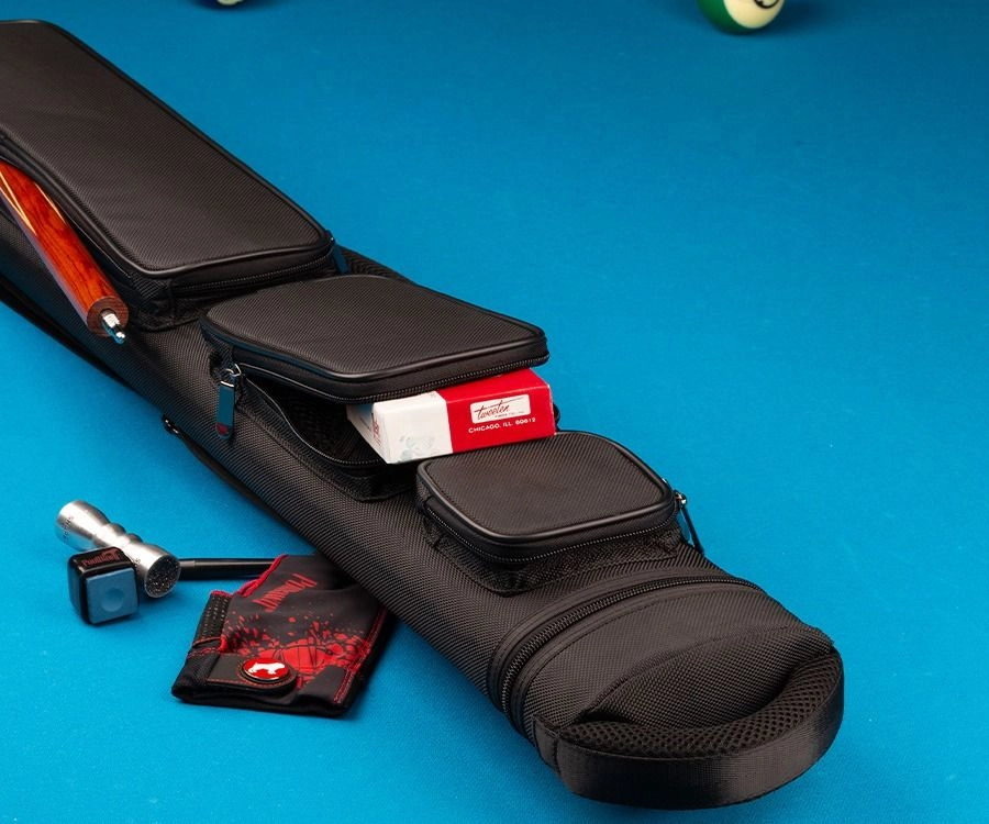 A black pool cue case on a blue pool table, shown with a wooden cue shaft tucked into one of its zippered compartments. A red and white box, a small black accessory, and a red-and-black card are placed beside the case.