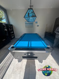 Pool table with blue felt in a room with a lamp and window.
