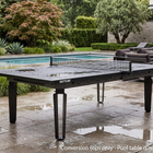 A black Kettler outdoor pool table with a net, positioned on a wet stone patio next to a swimming pool in a landscaped garden.