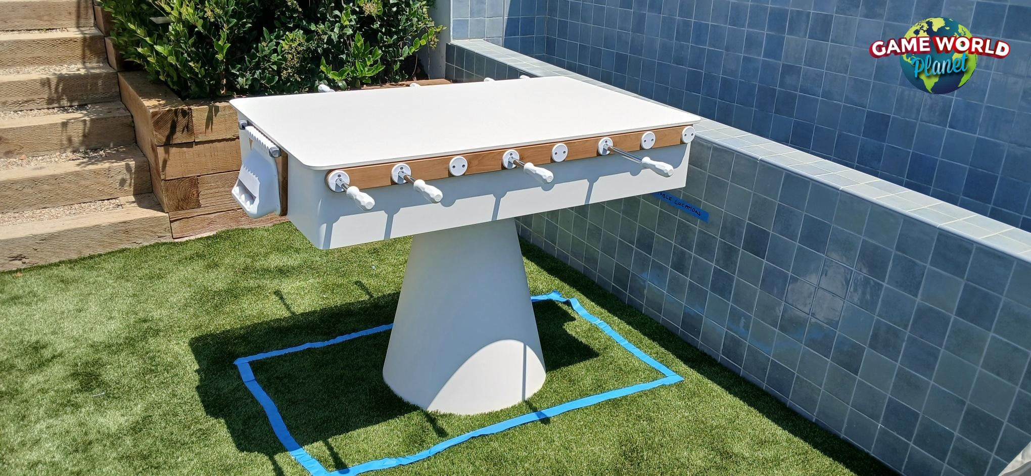 Kettler FAS Ciclope White Outdoor Foosball Table with cover and balls on grass near tiled wall.