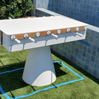 Kettler FAS Ciclope White Outdoor Foosball Table with cover and balls on grass near tiled wall.