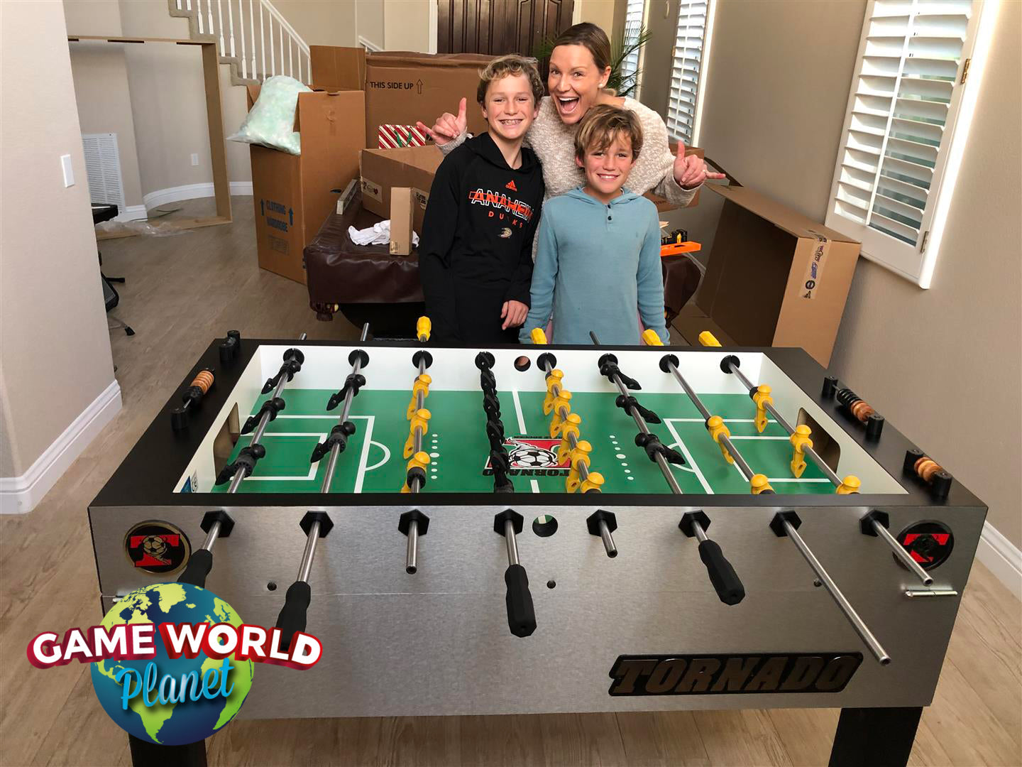 A woman and two children stand by a Tornado T-3000 Silver foosball table amid unpacked boxes.
