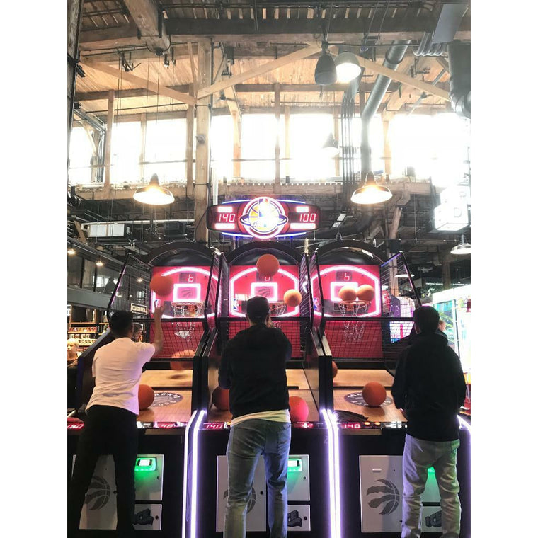 Three people play the ICE NBA GameTime Basketball Arcade Game under exposed beams and lights.