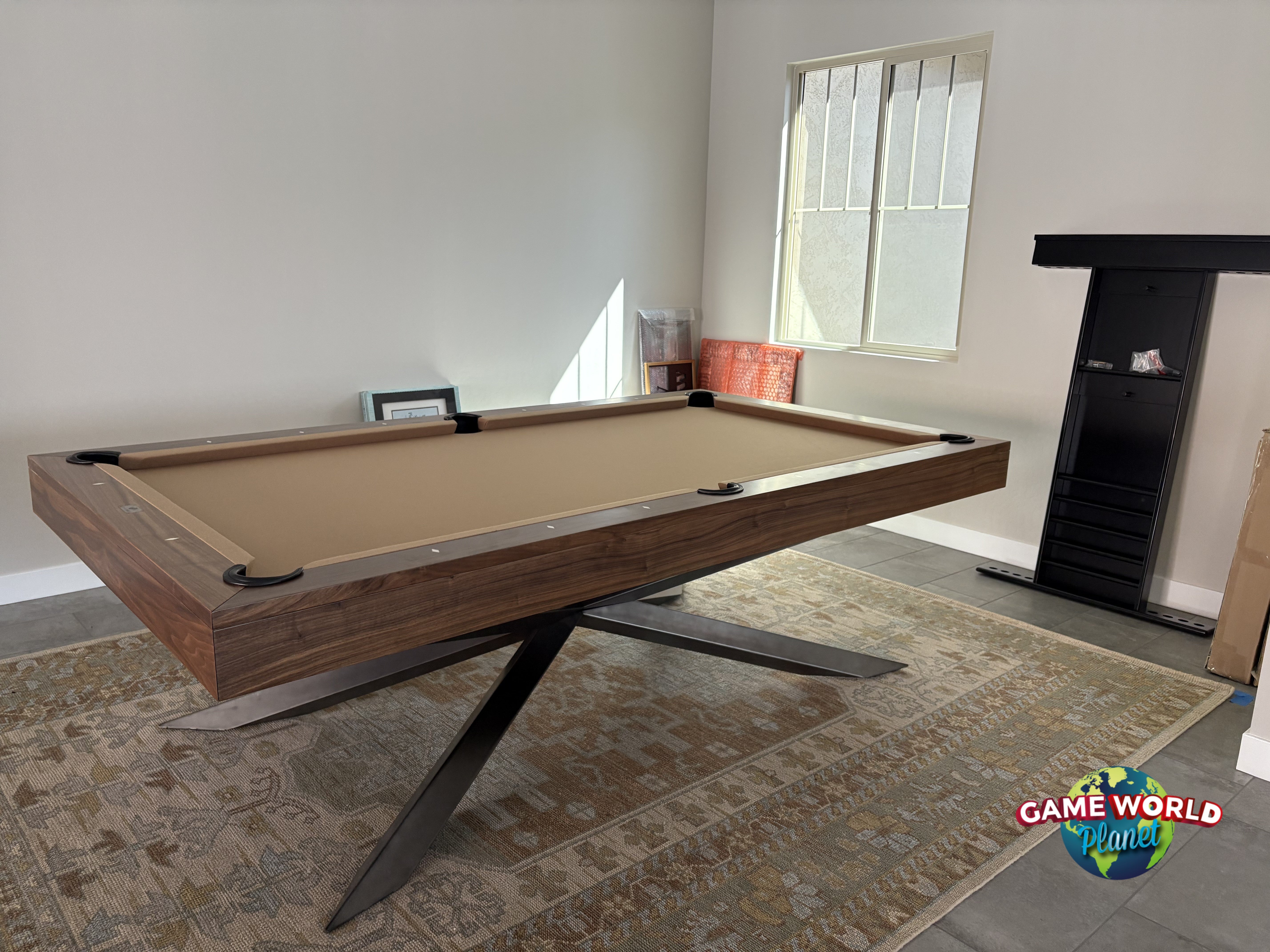 A modern pool table with a dark wood frame, tan felt surface, and black metal X-leg base, shown in a room with a patterned rug, fireplace, and window.