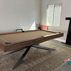 A modern pool table with a dark wood frame, tan felt surface, and black metal X-leg base, shown in a room with a patterned rug, fireplace, and window.