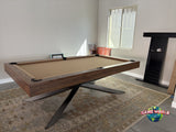 A modern pool table with a dark wood frame, tan felt surface, and black metal X-leg base, shown in a room with a patterned rug, fireplace, and window.