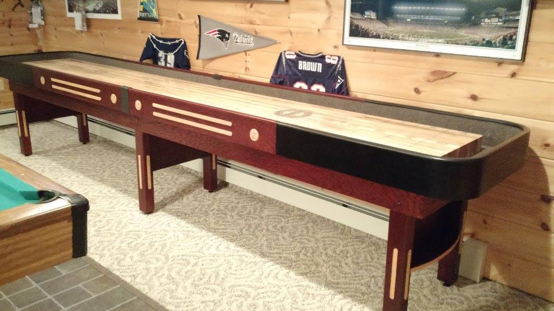 A Champion 9' The Grand Champion Shuffleboard Table in a sports-themed room with framed jerseys.