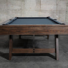 A Nixon Hunter 7' Slate Pool Table in brushed walnut stands before a concrete wall.
