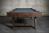 A Nixon Hunter 7' Slate Pool Table in brushed walnut stands before a concrete wall.