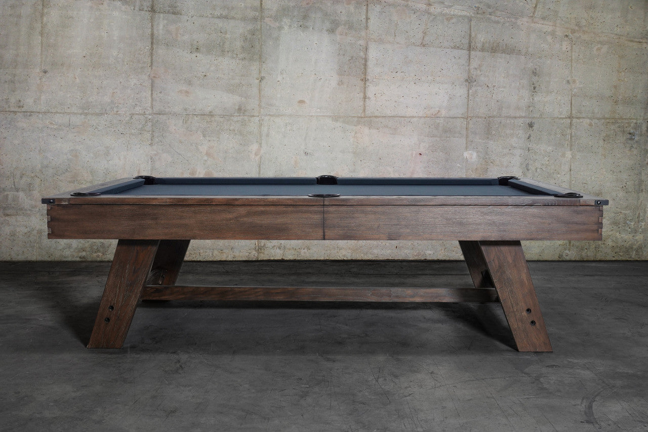 The Nixon Hunter 7' Slate Pool Table in Brushed Walnut by Nixon Billiards stands by a concrete wall.