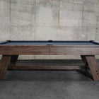 The Nixon Hunter 7' Slate Pool Table in Brushed Walnut by Nixon Billiards stands by a concrete wall.