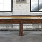 A Brunswick Canton 14' Shuffleboard Table in Black Forest sits before windows in a brick-walled room.