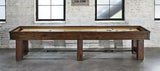 A Brunswick Canton 14' Shuffleboard Table in Black Forest sits before windows in a brick-walled room.