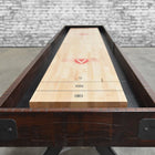 A Venture Williamsburg 12' Shuffleboard Table sits on concrete by an exposed brick wall.
