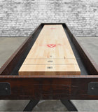 A Venture Williamsburg 12' Shuffleboard Table sits on concrete by an exposed brick wall.