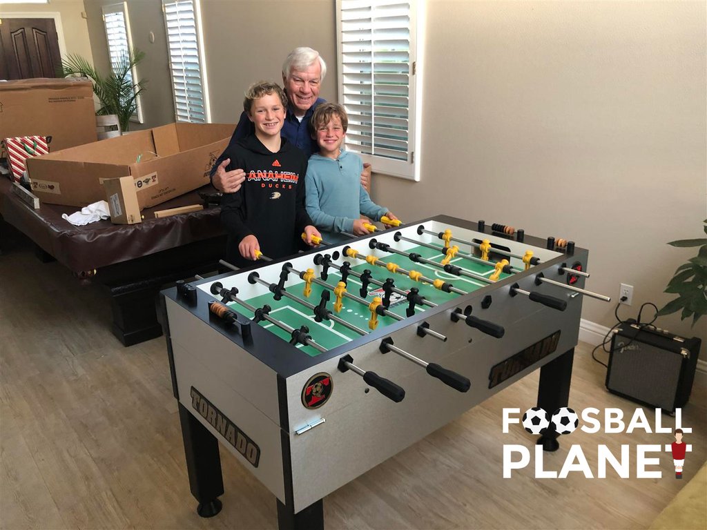 Three people behind a Tornado T-3000 Foosball Table in a living room with moving boxes.